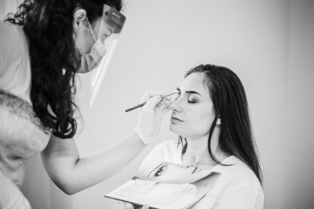 Truccatrice con visiera applica il trucco occhi a una donna in divisa durante il servizio fotografico Alta Clinic del 2020