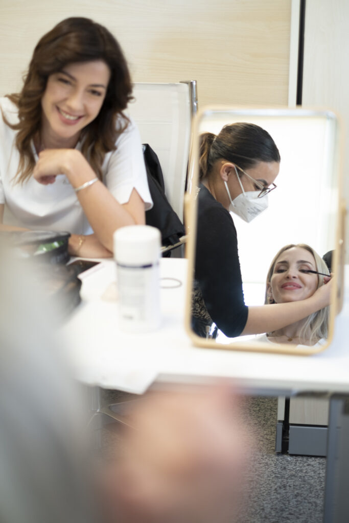 Donna sorridente al tavolo del trucco mentre lo specchio riflette la truccatrice con mascherina nel backstage 2022