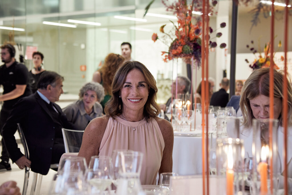 Ospite sorridente seduta a un tavolo con candele e fiori arancioni durante la cena della Cesvi Charity Night 2023