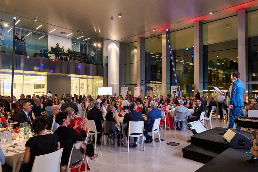 Presentatore sul palco e ospiti seduti ai tavoli nella grande sala vetrata della Cesvi Charity Night 2022